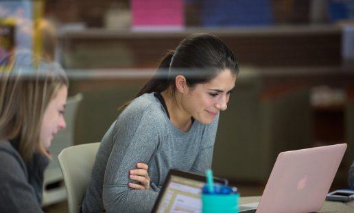 Two women sit at a table working on laptops on campus.