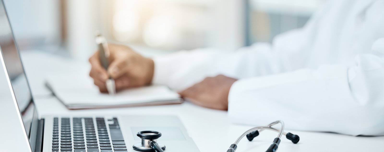 A human writing on a notepad with a laptop and stethoscope on the desk
